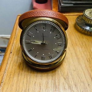 May include: A vintage travel alarm clock with a gold-tone case and a brown leather cover. The clock face is gray with black Roman numerals and hands. The clock reads "7 Jewels" and "Westclox".