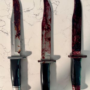 May include: Three stainless steel knives with black handles and silver accents. The blades are covered in a red substance, suggesting a horror or Halloween theme. The knives are arranged vertically.
