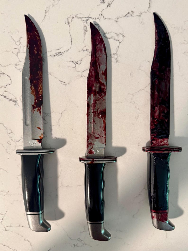 May include: Three stainless steel knives with black handles and silver accents. The blades are covered in a red substance, suggesting a horror or Halloween theme. The knives are arranged vertically.