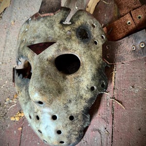 May include: A weathered, off-white hockey mask with a red triangle accent and numerous circular eye holes. The mask has a distressed appearance, with visible wear and tear, and is accompanied by a brown leather strap.