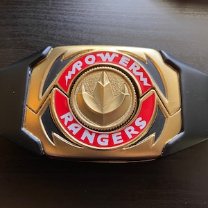 May include: A gold and black Power Rangers belt buckle with the words "POWER RANGERS" in red lettering. The center features a gold emblem. The buckle is a replica of the one used in the Power Rangers television series.