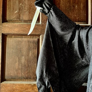 May include: A black costume sleeve and glove holding a silver knife with a light-colored blade. The costume is made of a dark, textured fabric. The background is a wooden door.