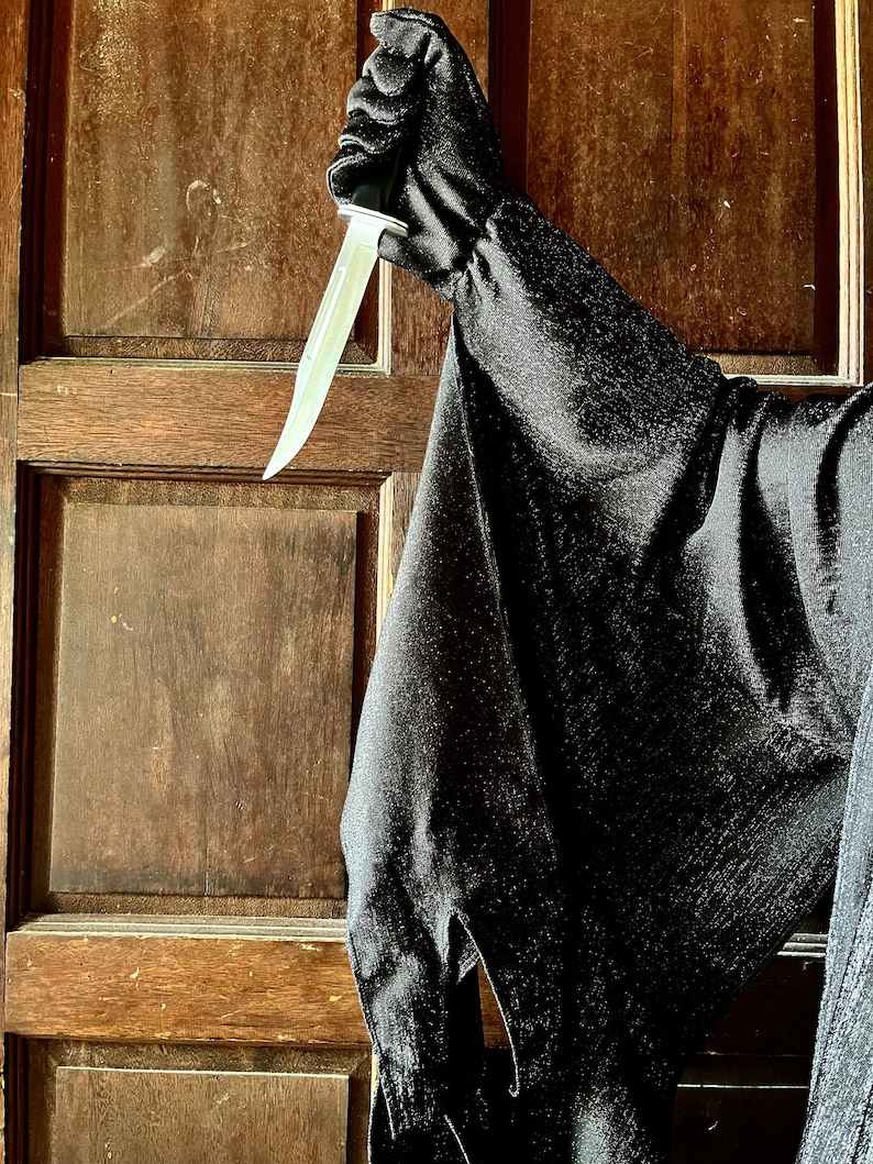 May include: A black costume sleeve and glove holding a silver knife with a light-colored blade. The costume is made of a dark, textured fabric. The background is a wooden door.