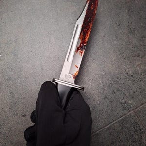 May include: A stainless steel knife with a curved blade, bearing the "BUCK USA" mark. The blade is stained with a dark red substance. The knife is held by a person wearing a black glove.