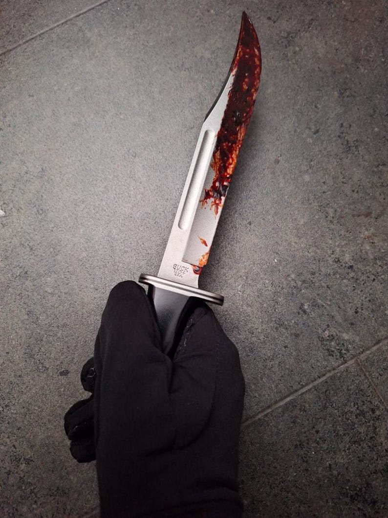 May include: A stainless steel knife with a curved blade, bearing the "BUCK USA" mark. The blade is stained with a dark red substance. The knife is held by a person wearing a black glove.