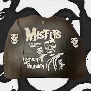 May include: A black long-sleeve shirt featuring a white graphic print of the Misfits logo, the text "evil never dies....." and "Legacy of Brutality," along with skeletal imagery and skull icons on both sleeves.