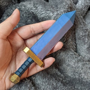 Pode incluir: Uma pequena espada de madeira com uma lâmina pintada de azul e roxo e uma empunhadura pintada de ouro. A espada está sendo segurada em uma mão.