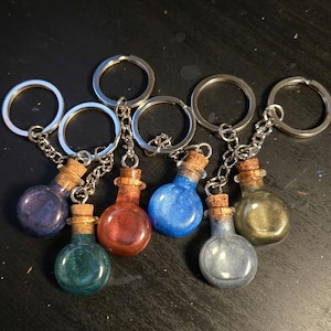 May include: A set of seven small glass bottles with cork stoppers, each filled with a different colored glitter. The bottles are attached to silver keychains.