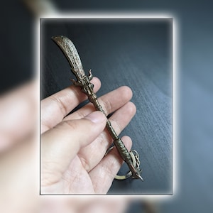 May include: A bronze-colored metal keychain shaped like a sword with intricate details. The sword has a curved blade and a handle wrapped in brown string.