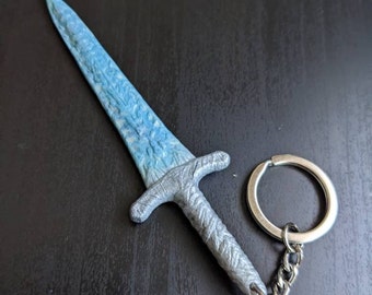 May include: A silver and blue sword keychain with a detailed design. The sword is attached to a silver key ring and chain.