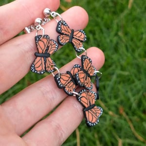 May include: A butterfly bracelet featuring orange and black monarch butterfly charms. The bracelet is made of silver-colored metal and has a delicate design. The butterflies have detailed wing patterns and are connected by small rings.
