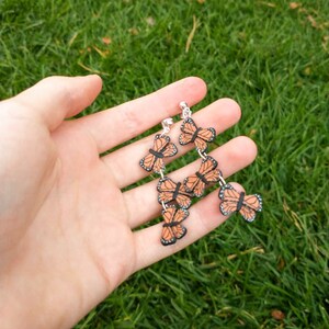 May include: Dangling earrings featuring a series of orange and black butterfly charms. The butterfly charms are connected by silver-colored chains. The earrings are held in a hand against a backdrop of green grass.