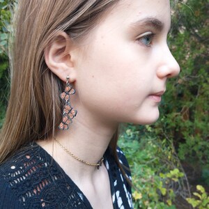 May include: Dangling earrings featuring a series of orange and black butterfly pendants. The earrings are suspended from a silver post and hang vertically. A gold chain necklace is also visible.