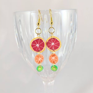 Puede incluir: Pendientes colgantes con diseño de rodajas de fruta. Cada pendiente tiene un gancho y una cadena dorados, con rodajas de pomelo rojo, naranja y lima. Los pendientes están hechos de arcilla polimérica.