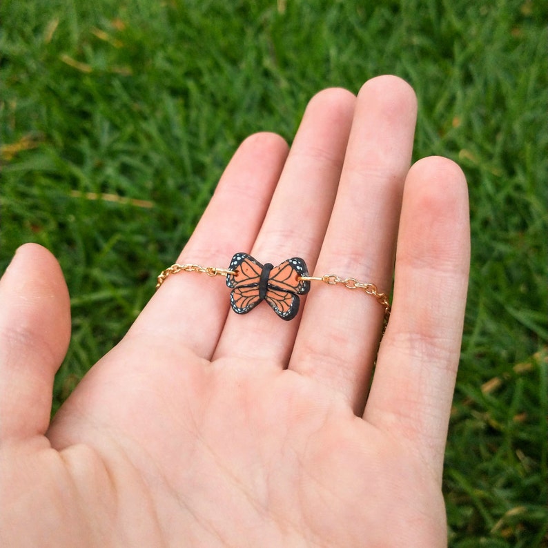 May include: A gold-colored chain bracelet featuring a monarch butterfly charm. The butterfly charm is orange, black, and white. The bracelet is displayed on a hand against a background of green grass.
