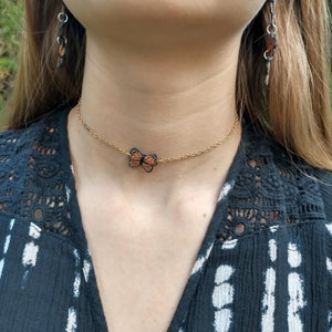 May include: A gold-colored chain necklace with a butterfly pendant. The butterfly is orange and black, with a delicate design. Matching butterfly earrings are also visible. The jewelry is set against a dark blue patterned top.