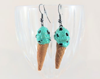 Puede incluir: Un par de pendientes novedosos con forma de conos de helado de menta con chispas de chocolate. Las bolas son de color verde claro con acentos de chispas de chocolate oscuro, sobre conos de gofre marrones. Los pendientes cuelgan de ganchos plateados.