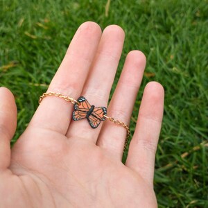 May include: A gold-colored chain bracelet featuring a monarch butterfly charm. The butterfly charm is orange, black, and white. The bracelet is displayed against a background of green grass.