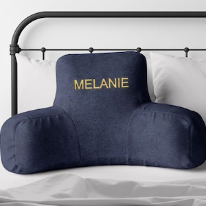 Puede incluir: Un coj&iacute;n de lectura azul marino con reposabrazos, bordado con el nombre "MELANIE" en hilo dorado. El coj&iacute;n est&aacute; sobre una cama con almohadas blancas y un cabecero de metal negro. El coj&iacute;n est&aacute; dise&ntilde;ado para soporte lumbar.