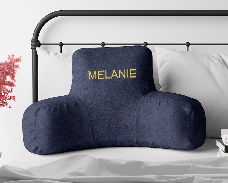 Puede incluir: Un coj&iacute;n de lectura azul marino con reposabrazos, bordado con el nombre "MELANIE" en hilo dorado. El coj&iacute;n est&aacute; sobre una cama con almohadas blancas y un cabecero de metal negro. El coj&iacute;n est&aacute; dise&ntilde;ado para soporte lumbar.