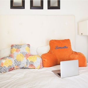 Puede incluir: Dos cojines de descanso para la cama sobre una cama. Uno es naranja con el nombre "Samantha" bordado en azul. El otro tiene dise&ntilde;os florales amarillos, grises y naranjas. Hay un port&aacute;til abierto en la cama.