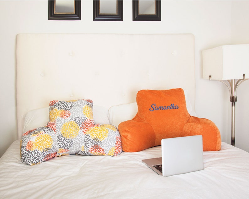 Puede incluir: Dos cojines de descanso para la cama sobre una cama. Uno es naranja con el nombre "Samantha" bordado en azul. El otro tiene dise&ntilde;os florales amarillos, grises y naranjas. Hay un port&aacute;til abierto en la cama.