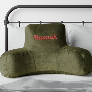 Puede incluir: Coj&iacute;n de descanso para cama verde oliva con el nombre "Hannah" bordado en rojo. La almohada tiene un dise&ntilde;o curvo con reposabrazos, colocada contra una almohada blanca y una estructura de cama de metal negro.