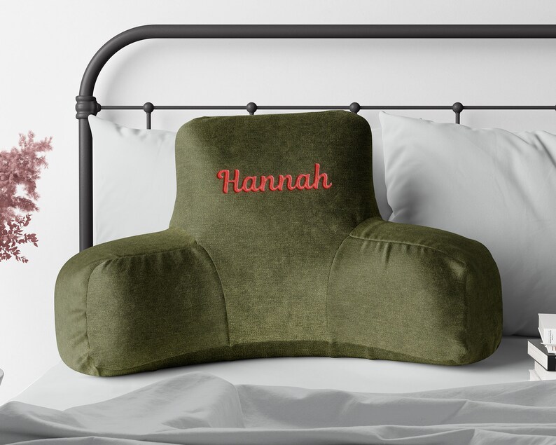 Puede incluir: Coj&iacute;n de descanso para cama verde oliva con el nombre "Hannah" bordado en rojo. La almohada tiene un dise&ntilde;o curvo con reposabrazos, colocada contra una almohada blanca y una estructura de cama de metal negro.