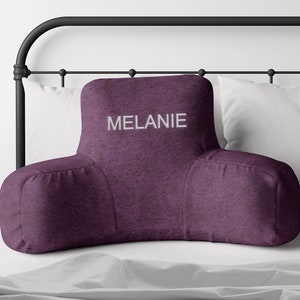 Puede incluir: Un coj&iacute;n de descanso para cama de color ciruela con reposabrazos, bordado con el nombre "MELANIE" en blanco. El coj&iacute;n est&aacute; sobre una cama con almohadas blancas y una cabecera de metal negro. El coj&iacute;n est&aacute; dise&ntilde;ado para soporte lumbar.