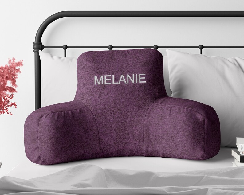 Puede incluir: Un coj&iacute;n de descanso para cama de color ciruela con reposabrazos, bordado con el nombre "MELANIE" en blanco. El coj&iacute;n est&aacute; sobre una cama con almohadas blancas y una cabecera de metal negro. El coj&iacute;n est&aacute; dise&ntilde;ado para soporte lumbar.