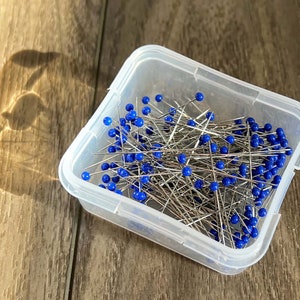 May include: A clear plastic container filled with sewing pins. The pins have silver metal shafts and blue plastic heads.