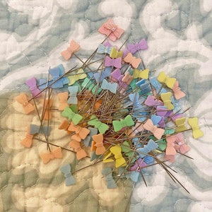 May include: A large collection of colorful bow-shaped pins with silver needles. The pins are arranged in a random pattern on a light-colored fabric background.
