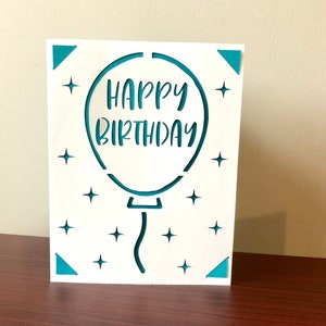 Op de afbeelding: Witte kaart met een uitgestanste turquoise ballon. De ballon heeft de woorden "Happy Birthday" erin. De kaart heeft kleine turquoise sterren rond de ballon.