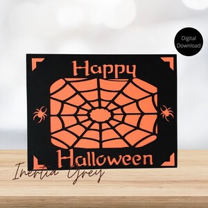 Puede incluir: Una tarjeta negra con una telaraña naranja recortada y las palabras "Happy Halloween" en blanco. La tarjeta está sobre una superficie de madera.