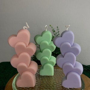 Könnte beinhalten: Drei herzförmige Kerzen in Pastellfarben: Rosa, Grün und Lavendel. Jede Kerze besteht aus gestapelten Herzen mit einem weißen Docht. Die Kerzen stehen auf einer Glasplatte auf einem Holzständer, mit grünem Hintergrund.