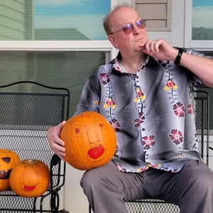 Può includere: Un uomo con una camicia a fiori grigia e rossa è seduto su una sedia di metallo nero tenendo in mano una zucca intagliata. Altre due zucche con facce dipinte sono sedute sulla sedia accanto a lui.