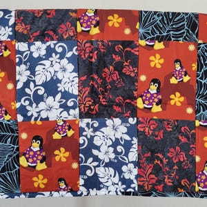May include: A patchwork quilt with a Hawaiian theme featuring penguins wearing purple shirts and flowers. The quilt is made of red, blue, black, and white fabric.