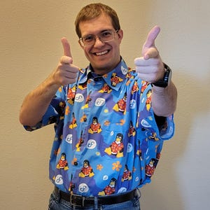 Op de afbeelding: Een man draagt een blauw, kort mouwig overhemd met een patroon van pinguïns en de letter 'F'. Hij glimlacht en wijst met beide handen.