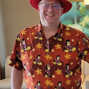 Può includere: Un uomo che indossa un cappello rosso e una camicia a maniche corte di colore arancione brillante con un motivo di pinguini che indossano camicie floreali viola e bianche. La camicia ha fiori gialli e accenti marroni.