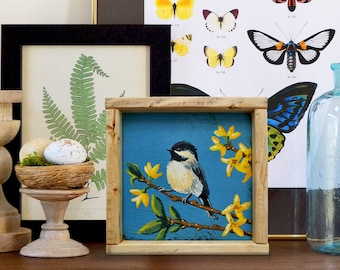 May include: A framed painting of a black-capped chickadee perched on a branch with yellow blossoms against a blue background.