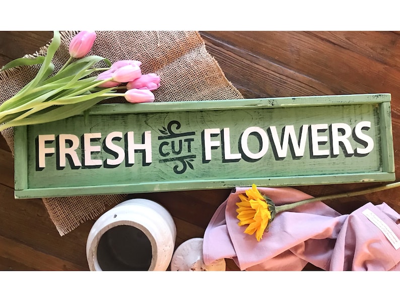 May include: A green wooden sign with white lettering that reads "FRESH CUT FLOWERS". The sign is sitting on a wooden surface with a burlap fabric and pink tulips in the background.
