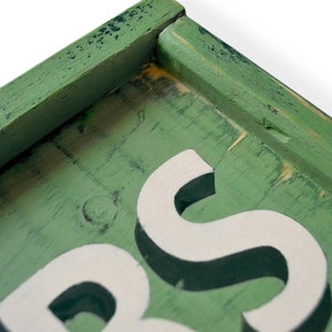 May include: Close-up of a green wooden sign with white raised letters spelling "S" and part of the letter "B". The sign has a distressed, vintage look.