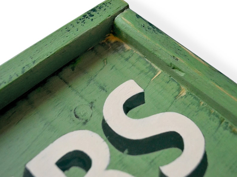 May include: Close-up of a green wooden sign with white raised letters spelling "S" and part of the letter "B". The sign has a distressed, vintage look.