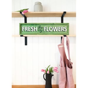 May include: A green wooden sign with white text that reads "FRESH CUT FLOWERS" hanging on a white wall with two wooden shelves. A pink apron hangs on a hook below the sign.