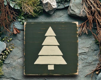 May include: A square, dark green wooden sign with a cream-colored Christmas tree design. The sign has a distressed, rustic look and is surrounded by natural elements like rocks, moss, and branches. A festive, seasonal decoration.