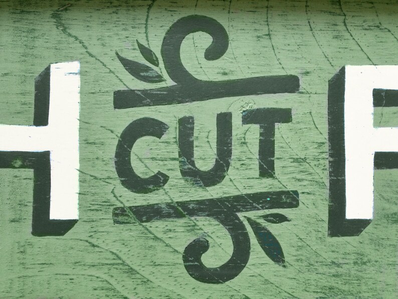May include: A green wooden sign with white and black painted letters that read "H CUT F". The letters are stylized and have a whimsical design.