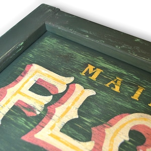 May include: A green wooden sign with a distressed finish. The sign has the words "MAIN" and "FLO" painted in gold and red letters.