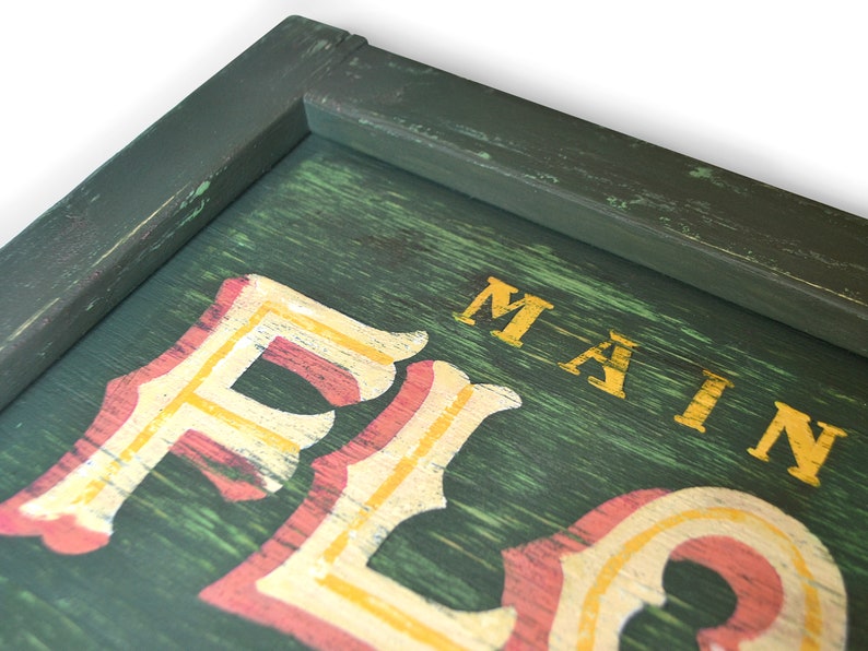 May include: A green wooden sign with a distressed finish. The sign has the words "MAIN" and "FLO" painted in gold and red letters.