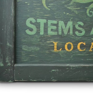 May include: A green wooden sign with white painted text that reads "STEMS AND LOCAL" in a vintage style font. The sign has a dark green frame.