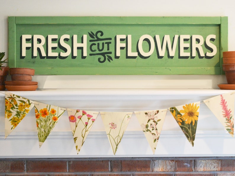 May include: A green sign with white lettering that reads "FRESH CUT FLOWERS" hangs above a white mantel. A floral garland with triangular flags featuring different flower illustrations is draped across the mantel.